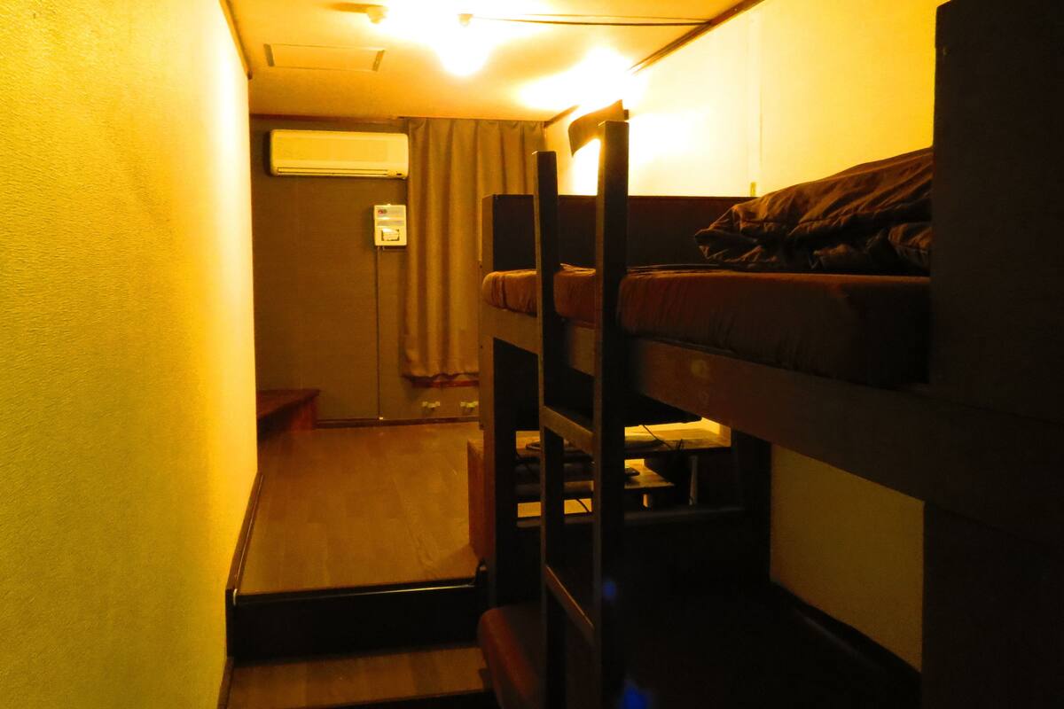 A narrow, dimly lit room with bunk beds, a small table, and a window with curtains.