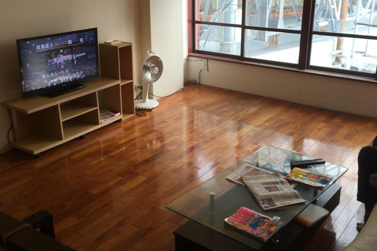 A living room with a television on a wooden stand, a glass coffee table with magazines, and a window with a view of construction outside.
