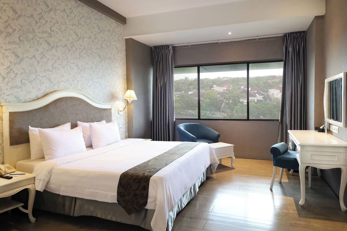 Habitación de hotel con una cama grande, un escritorio, una silla y vistas a la ciudad a través del ventanal.