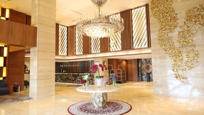 Een lobby met een marmeren vloer, een centraal hanglamp, een receptiebalie en een decoratieve muur met bladgoud patronen.