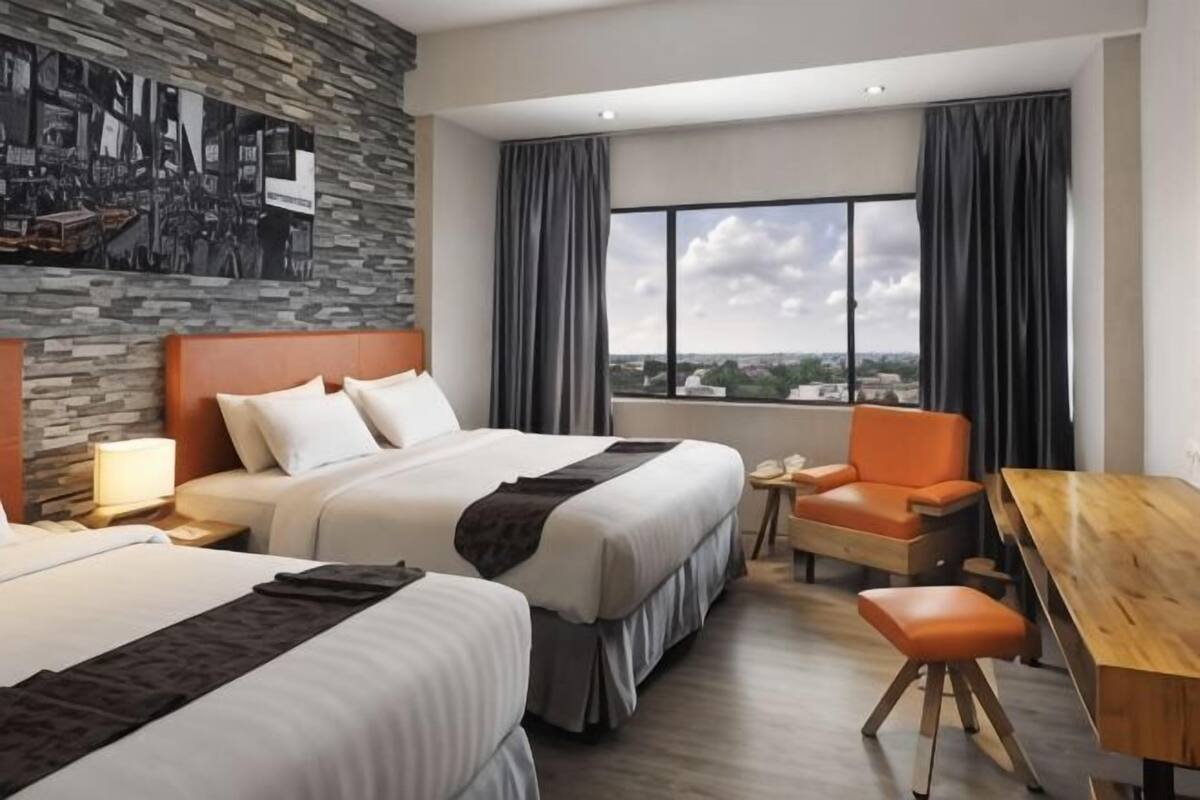 Una habitación de hotel con dos camas, un escritorio, una silla, un ventanal con cortinas y vistas a la ciudad.