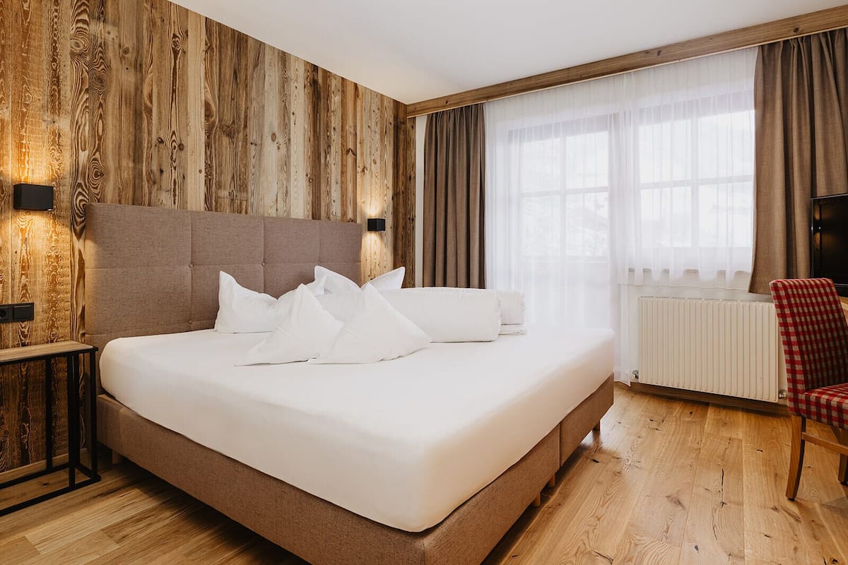 A hotel room with a large bed, wooden panel walls, a radiator, a red checkered chair, and a window with curtains.