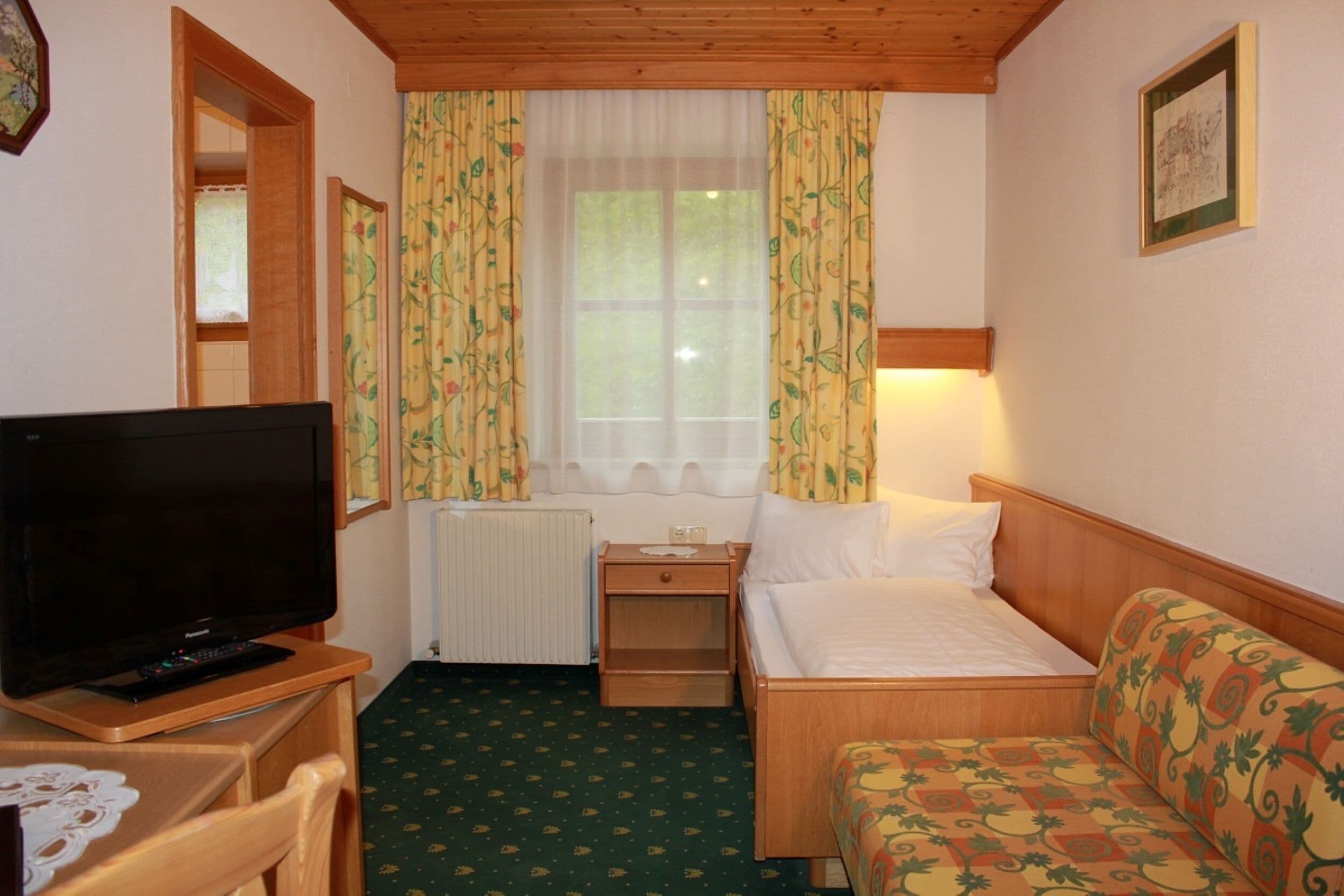A small, cozy room with a bed, a TV, a nightstand, and a window with curtains.