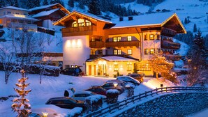 A multi-story hotel with a snow-covered exterior, a parking area with several cars, and a mountainous backdrop. - Hotel Dorfer (Grossarl)