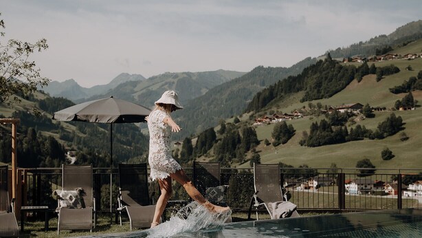 A person in a white dress walking on a wet surface near a pool with a mountainous backdrop. - Hotel Dorfer (Grossarl)