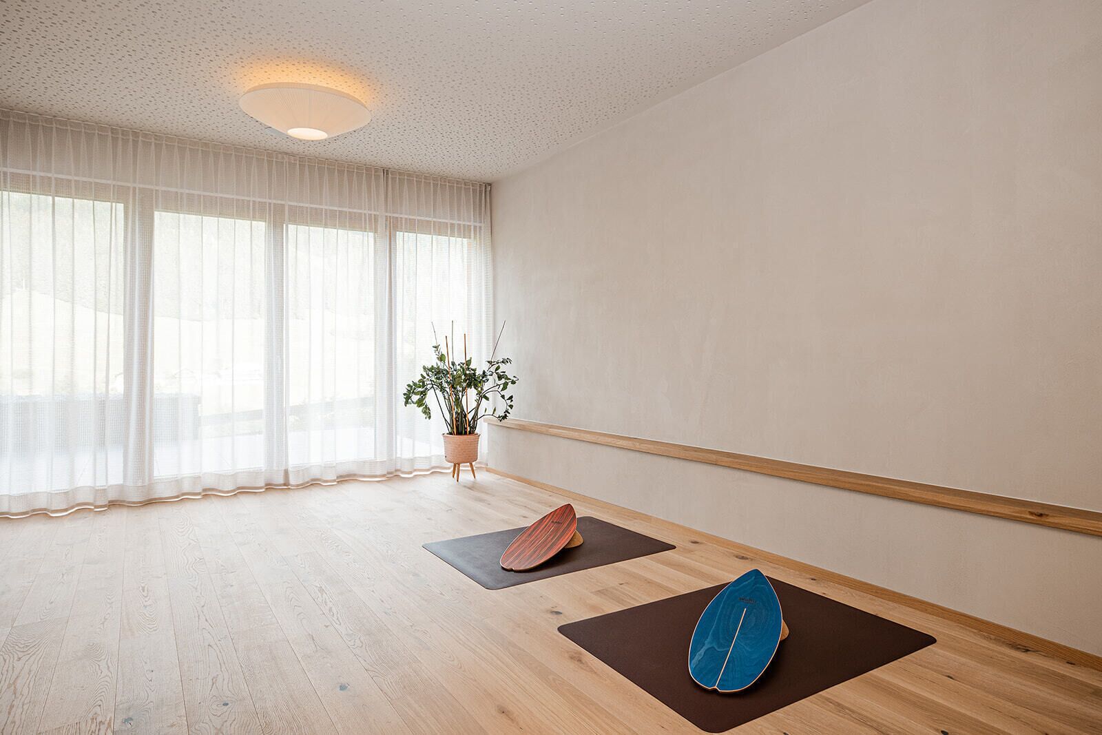 Quarto espaçoso com piso de madeira, uma janela grande com cortinas leves, uma planta em vaso e dois tapetes de yoga.