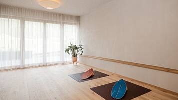Une pièce spacieuse avec un plancher en bois, une grande fenêtre ornée de rideaux légers, une plante en pot et deux tapis de yoga.