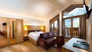 A hotel room with a bed, a desk, a chair, a TV, and a view of mountains through large windows. - Hotel Dorfer (Grossarl)