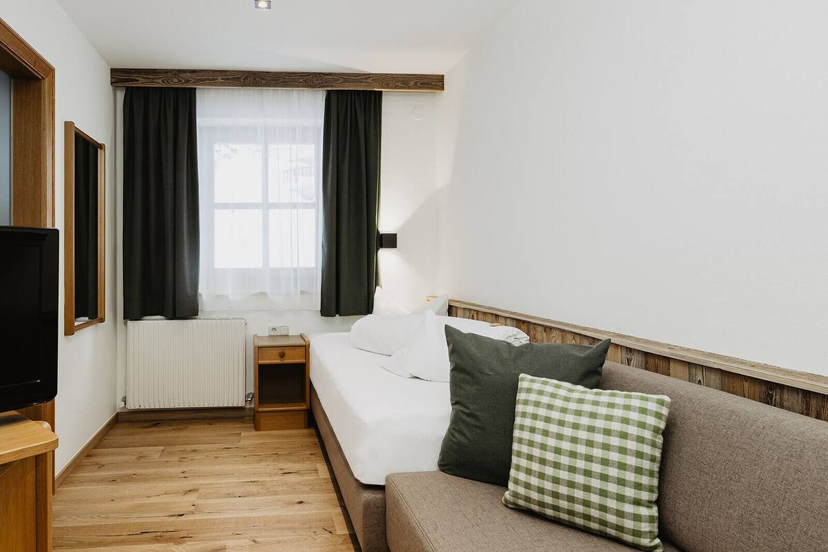 A hotel room with a bed, a wooden nightstand, a television, a window with curtains, and a radiator.