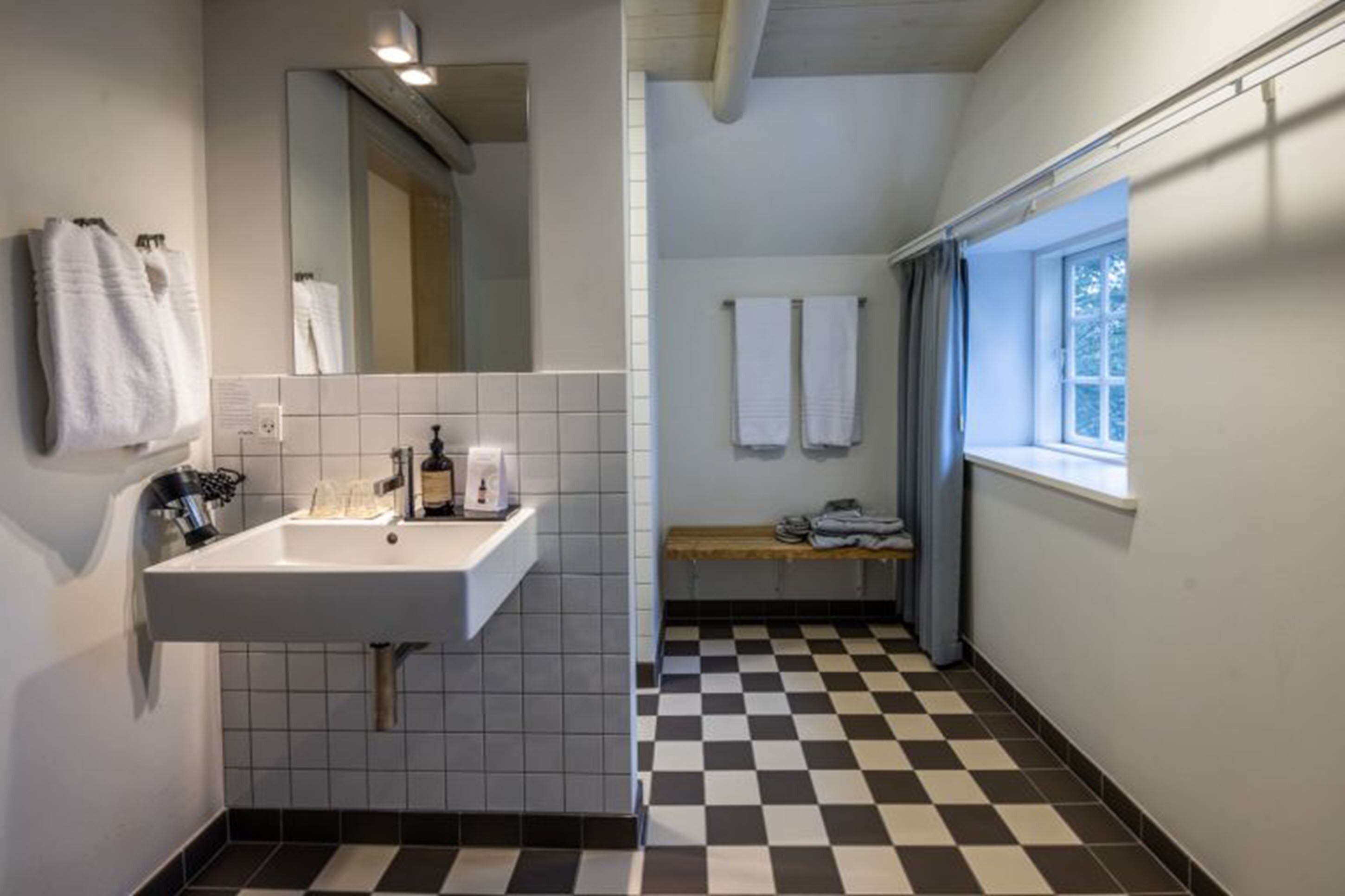 A bathroom with a white sink, mirror, and tiled floor. There are towels hanging on the wall and a bench with towels.