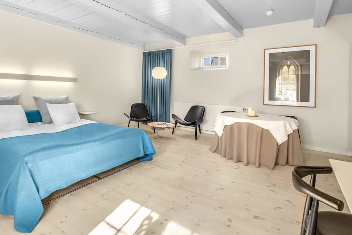 Une chambre d’hôtel moderne avec un grand lit, deux fauteuils, une petite table et une grande fenêtre avec des rideaux.