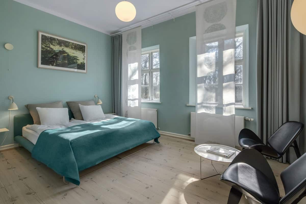 Une chambre à coucher avec un lit, des lampes de chevet, une chaise, une petite table et une fenêtre avec des rideaux.
