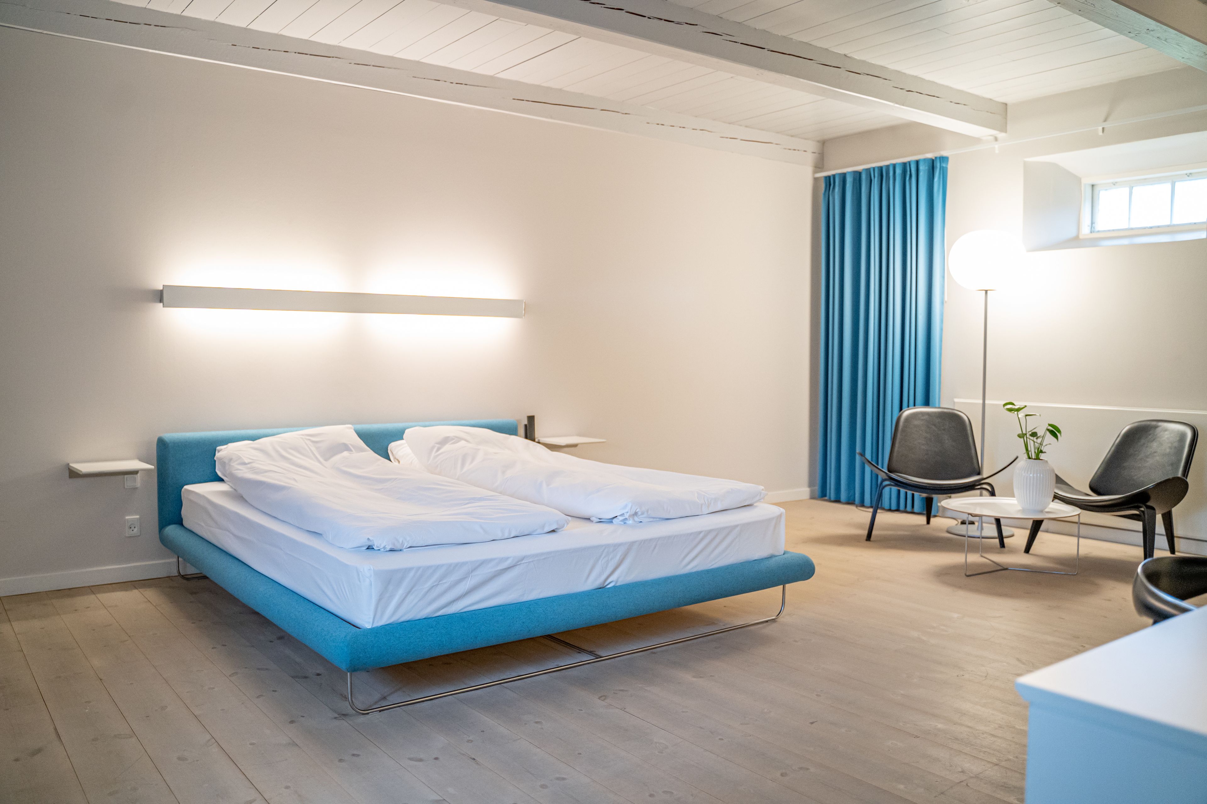 A modern hotel room with a blue bed, a bedside table, a floor lamp, and two chairs.