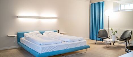 A modern hotel room with a blue bed, a bedside table, a floor lamp, and two chairs.