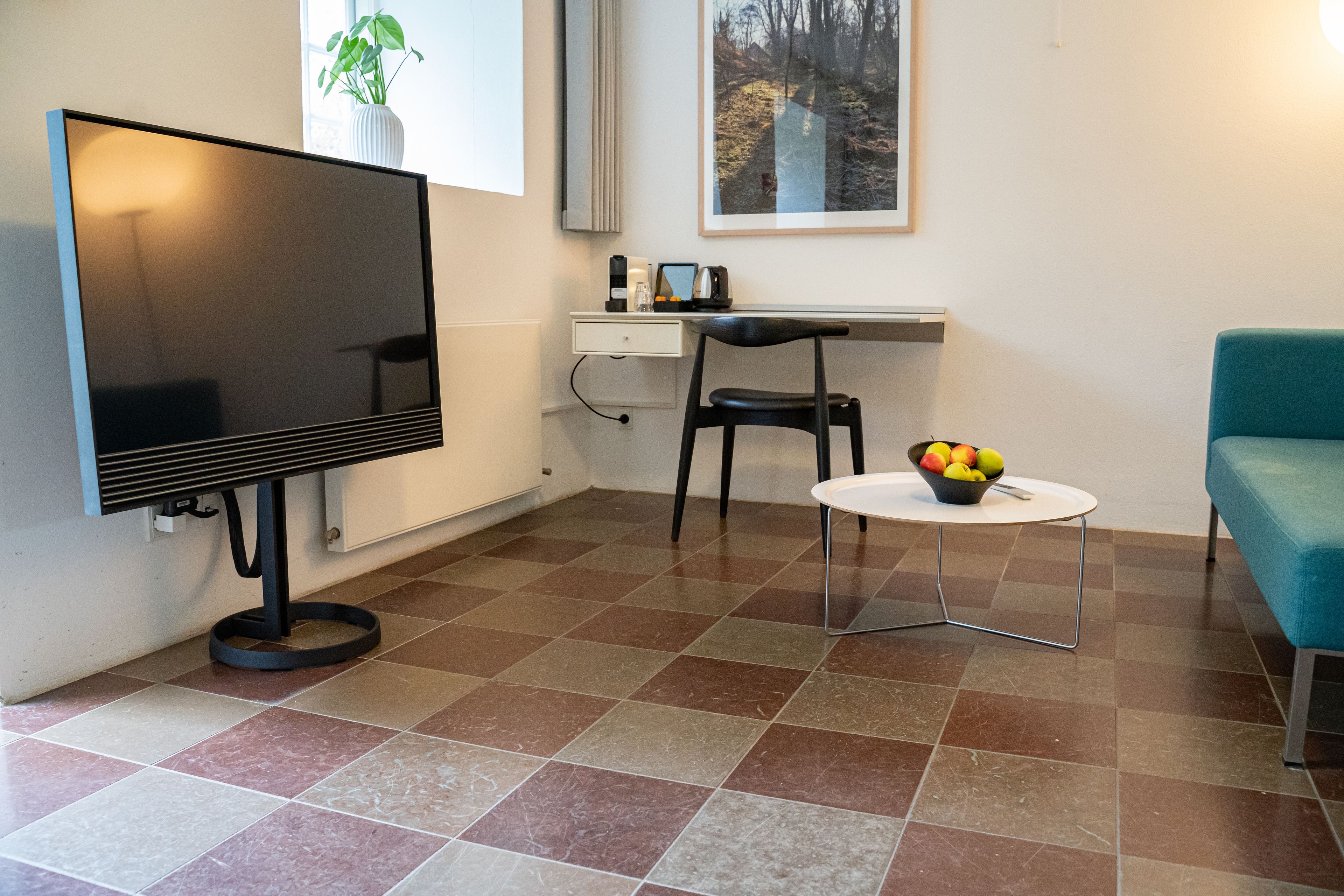 A modern living room with a flat-screen TV, a desk, a chair, a small round table with fruit, and a teal sofa.