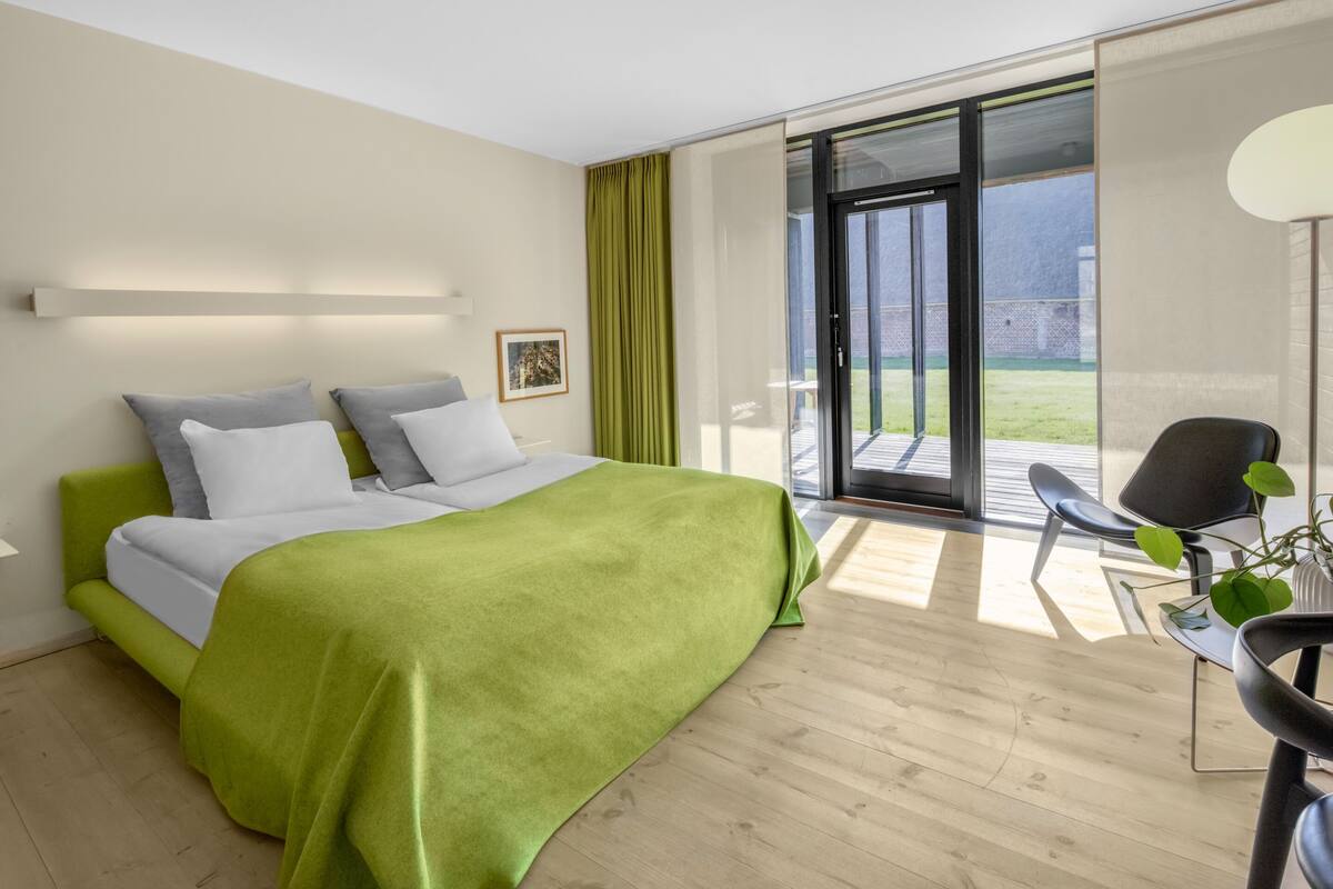 Une chambre moderne avec un grand lit, une couverture verte et une vue sur l’extérieur grâce à une porte-fenêtre coulissante.
