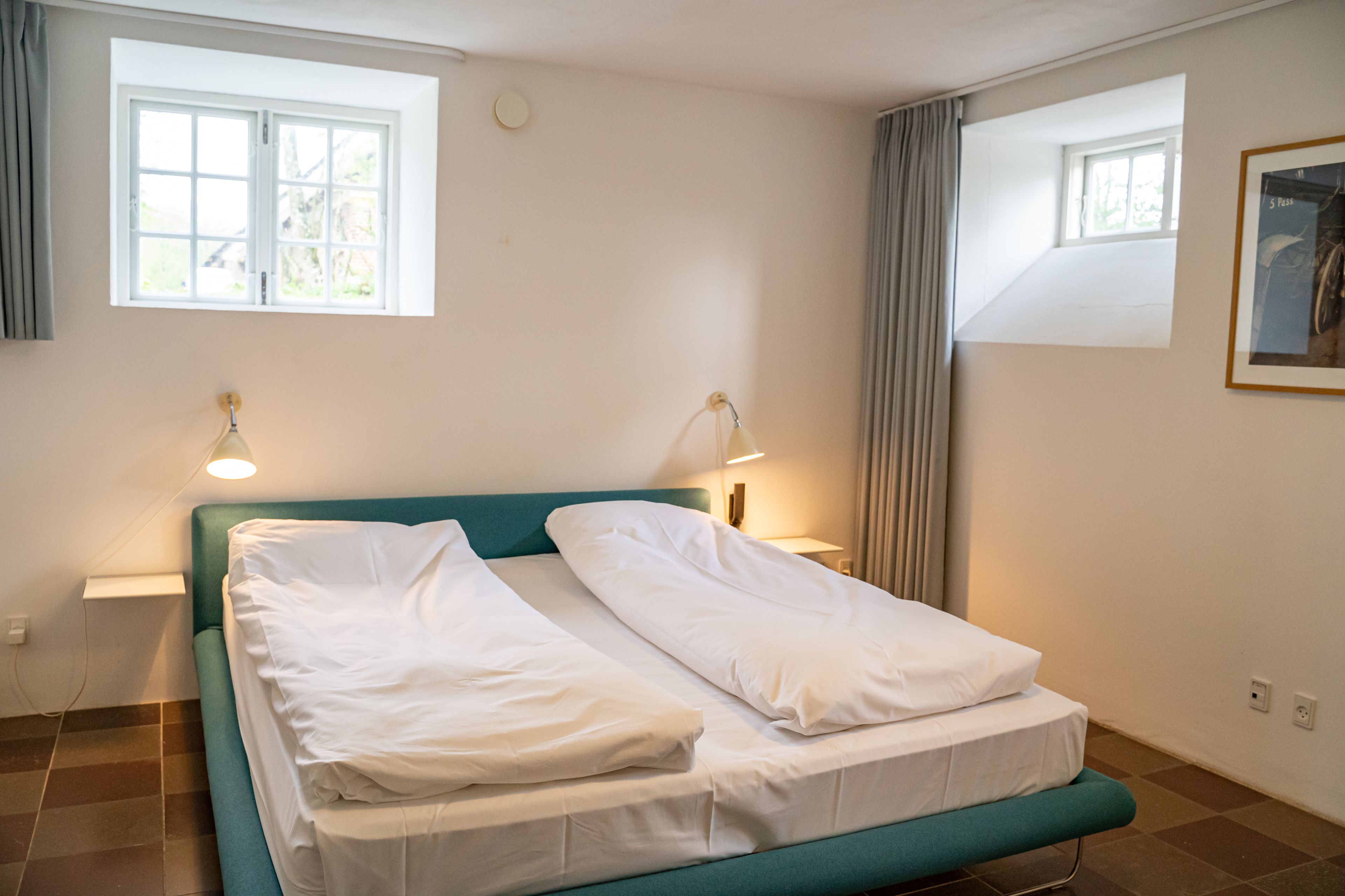 A neatly made bed with white linens in a room with a teal headboard, two bedside lamps, and a window with curtains.