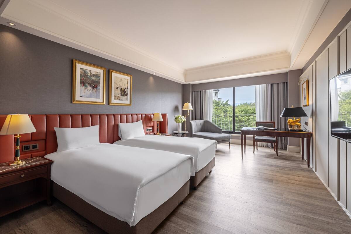 A hotel room with two beds, a desk, a chair, and a large window with a view of greenery.