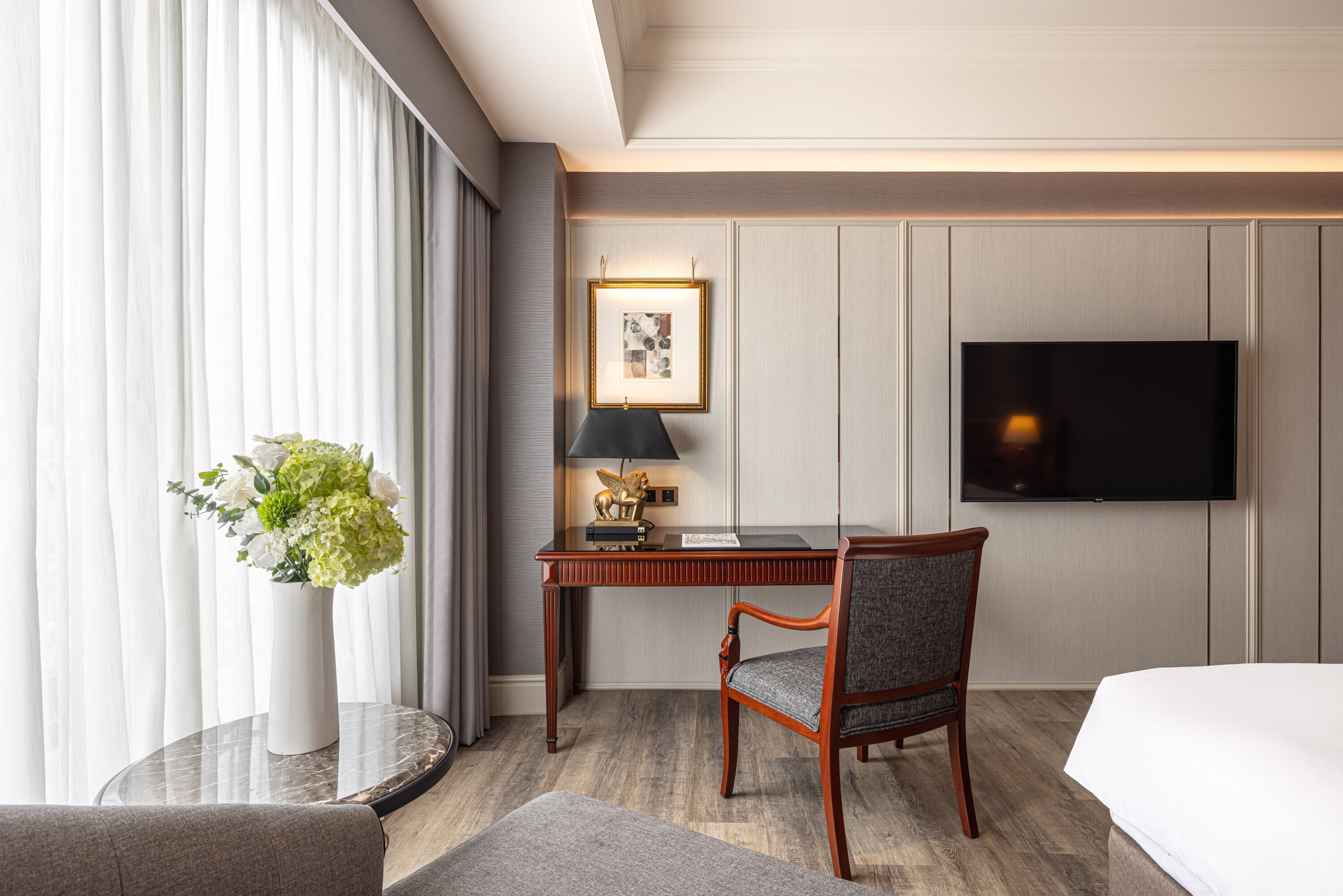 Habitación de hotel con cama, escritorio con silla, televisor, lámpara y un florero con flores.