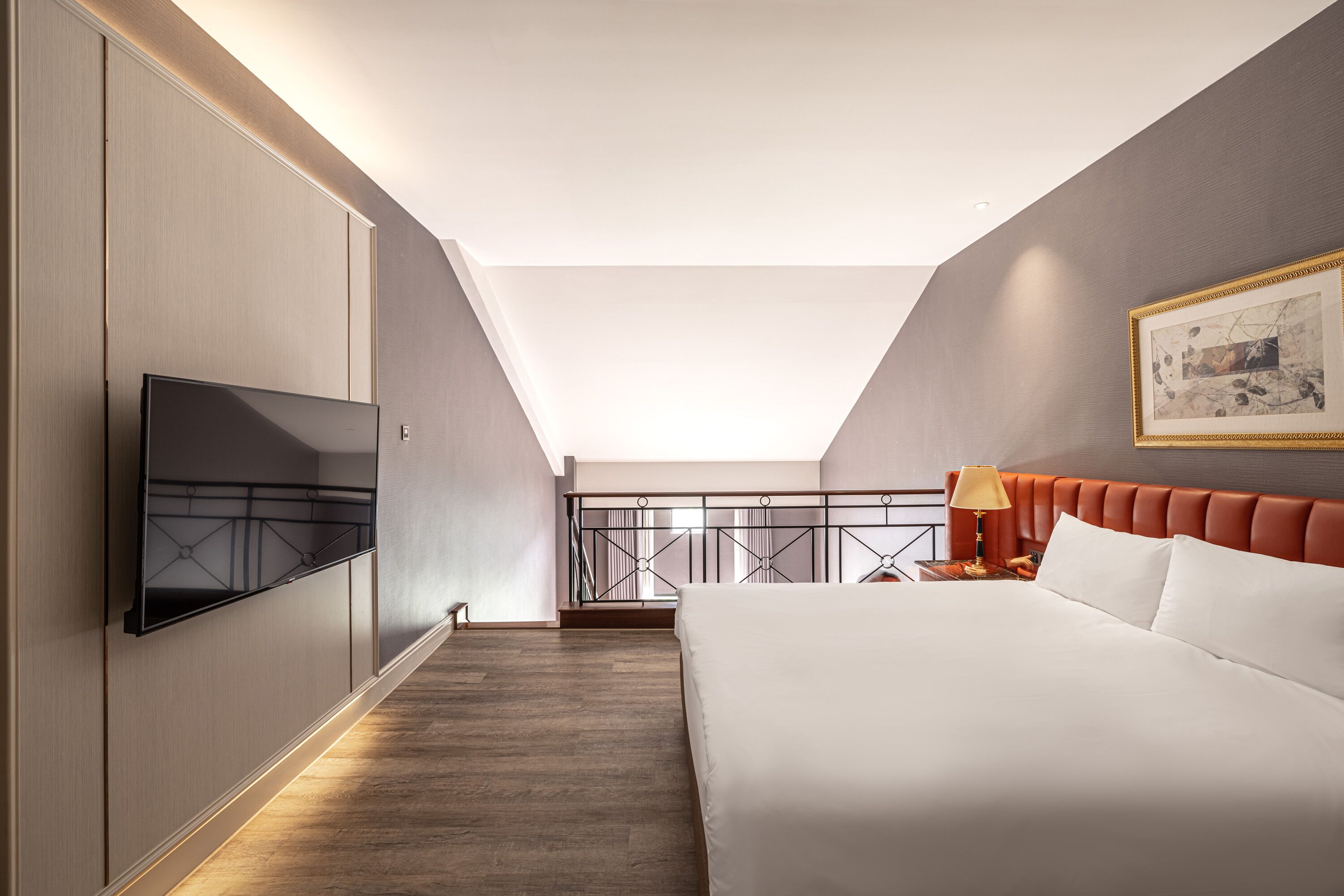 A modern hotel room with a large bed, a flat-screen TV mounted on the wall, a balcony with a railing, and a painting on the wall.