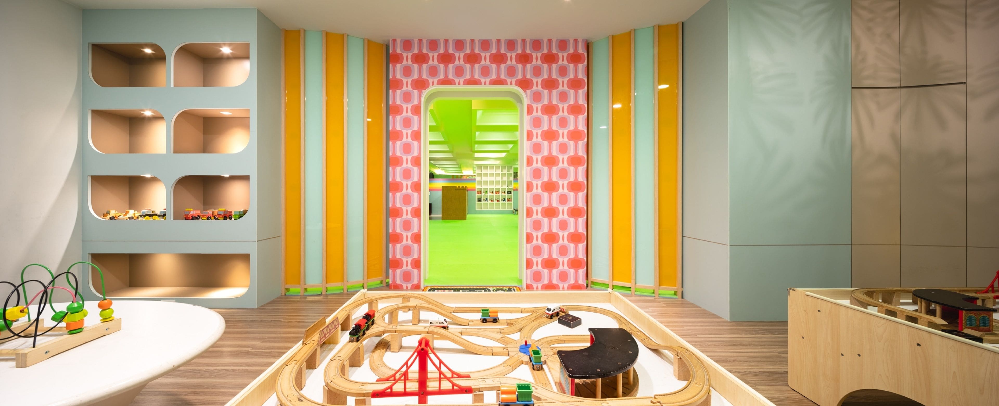 A playroom with a wooden train track layout, shelves with toys, and a colorful wall pattern.