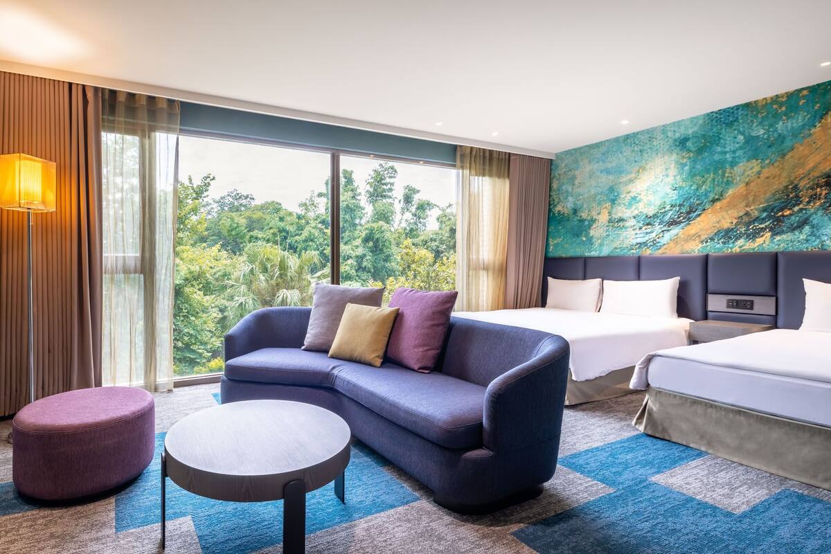 A modern hotel room with a large bed, a sofa, a small round table, and a view of the outdoors.