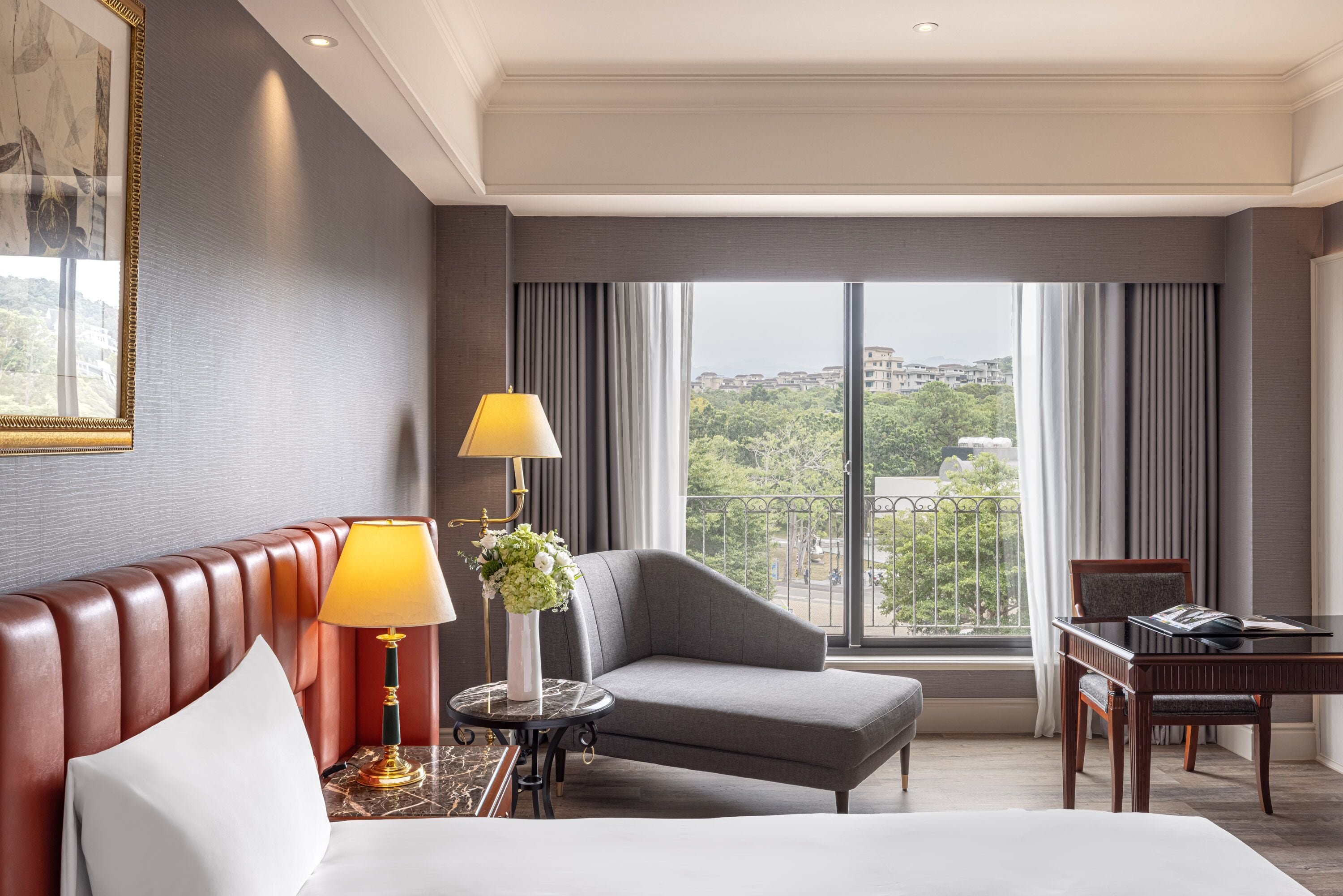 Habitación de hotel con una cama, un sillón gris, una mesita de noche con lámpara y vistas de la ciudad a través de ventanas amplias.