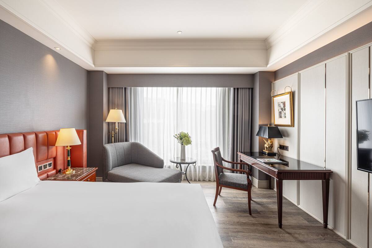 A hotel room with a large bed, a desk, two chairs, a TV, and a window with curtains.
