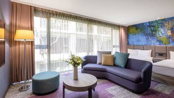 A modern hotel room with a sofa, a round coffee table, a bedside table, and a large window with curtains.
