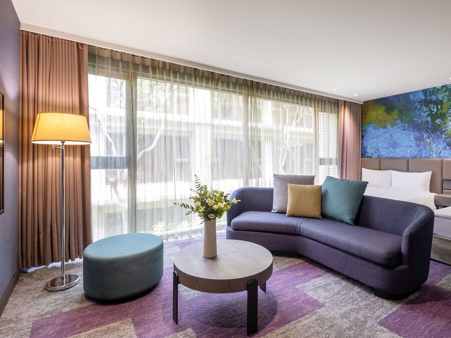 A modern hotel room with a sofa, a round coffee table, a bedside table, and a large window with curtains.
