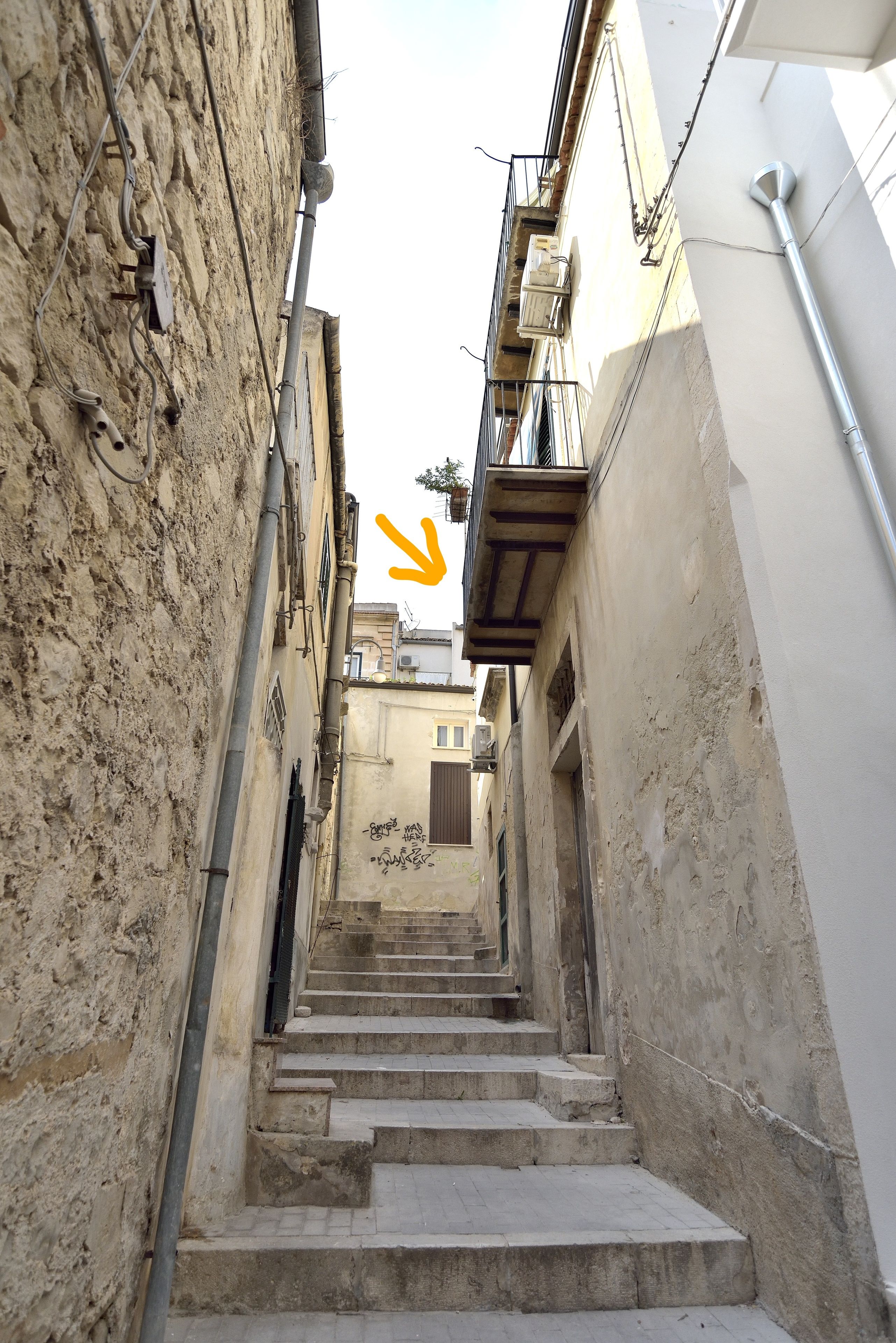 Um beco estreito com uma escadaria que sobe, ladeado por edifícios com canos e varandas à mostra.