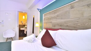 Habitación de hotel moderna con una cama bien hecha, una silla blanca y un baño visible a través de una puerta abierta.