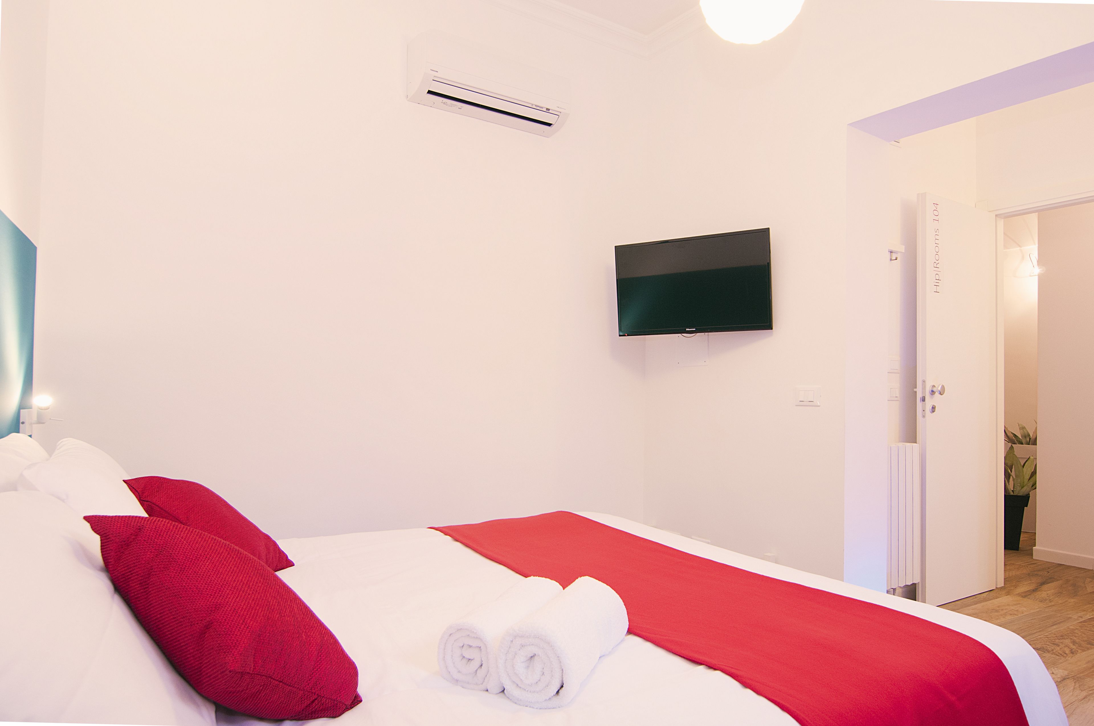 A hotel room with a bed, white bedding, red pillows, a red blanket, a flat-screen TV mounted on the wall, and a door leading to another room.