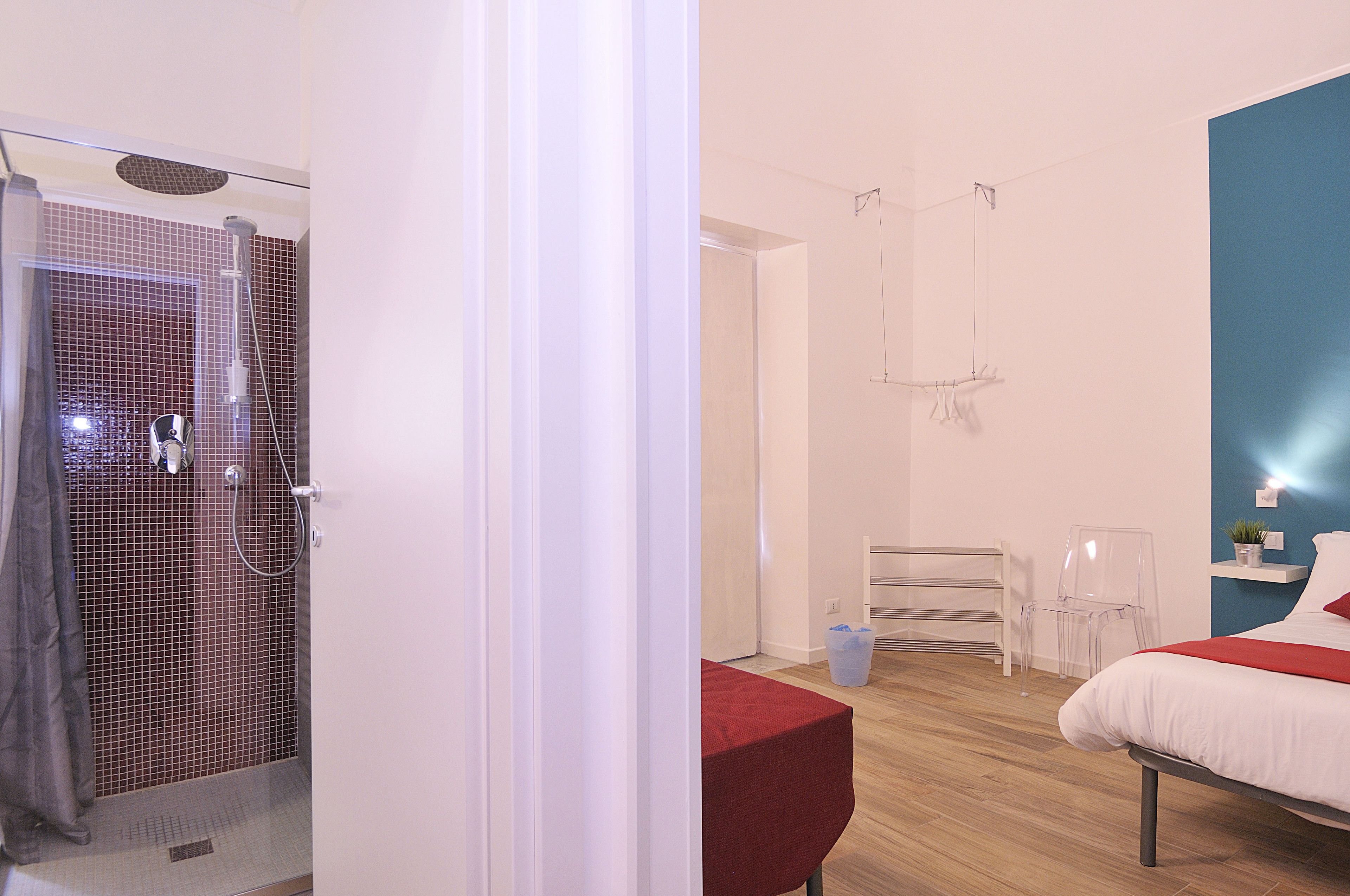 Un baño moderno con ducha, inodoro y lavabo. Hay una cama con una manta roja y una mesita de noche.