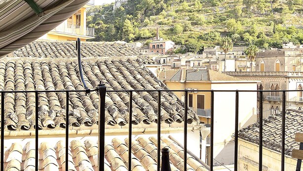 Un balcón con una mesa de madera, una botella de vino y una copa de vino. La vista muestra tejas de terracota y una colina con edificios y árboles.