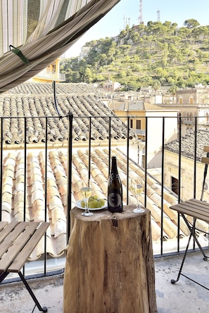 A balcony with a wooden table, a bottle of wine, and a glass of wine. The view includes terracotta roof tiles and a hillside with buildings and trees. - Modica Old Town Rooms (Modica)