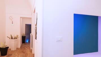 A hallway with a large blue artwork on the right wall, potted plants on the left, and a radiator in the center.