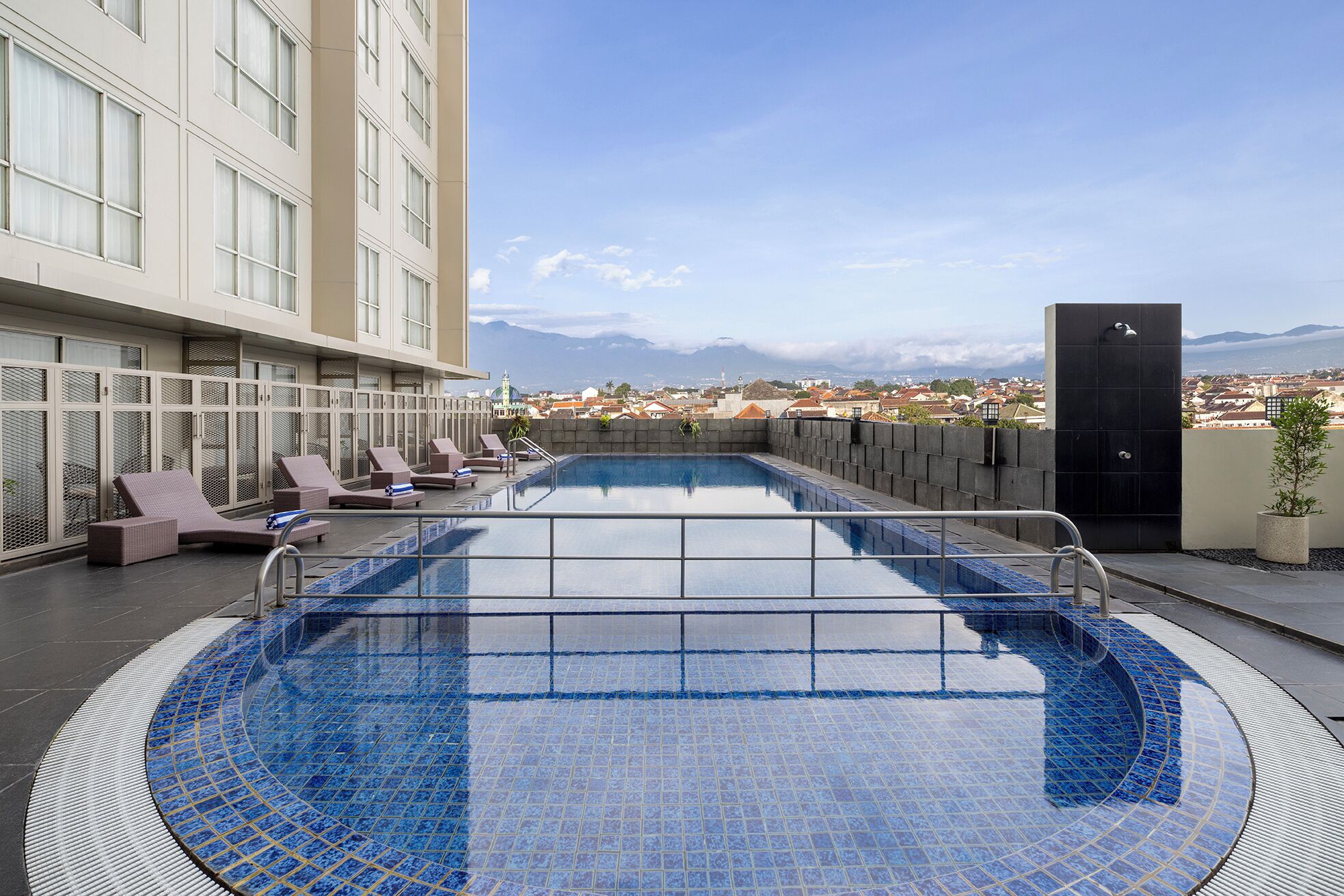 A rooftop pool with lounge chairs, a view of the city and mountains, and a modern building facade.