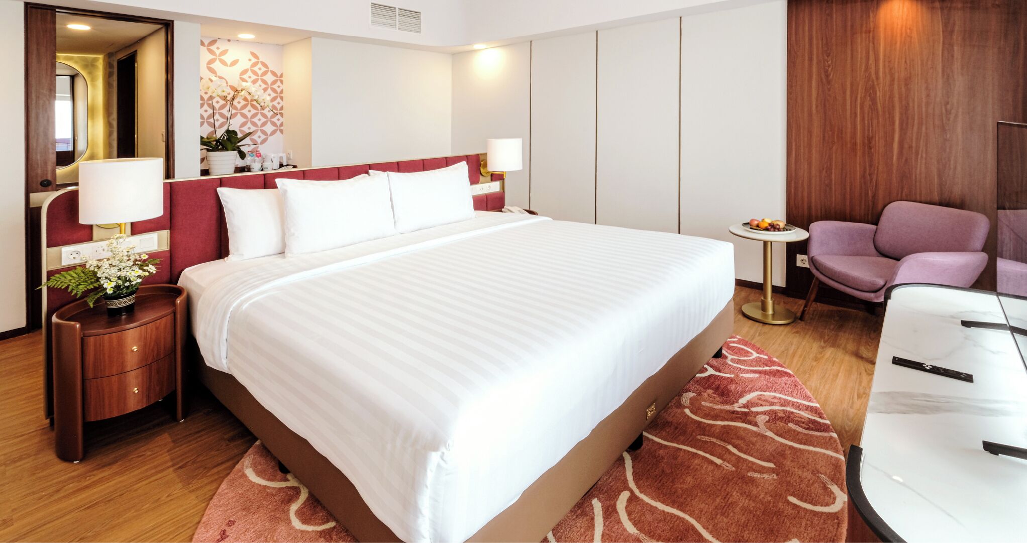 A hotel room with a large bed, a bedside table with a lamp, a small table with fruit, and a purple chair.