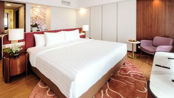 A hotel room with a large bed, a bedside table with a lamp, a small table with fruit, and a purple chair.