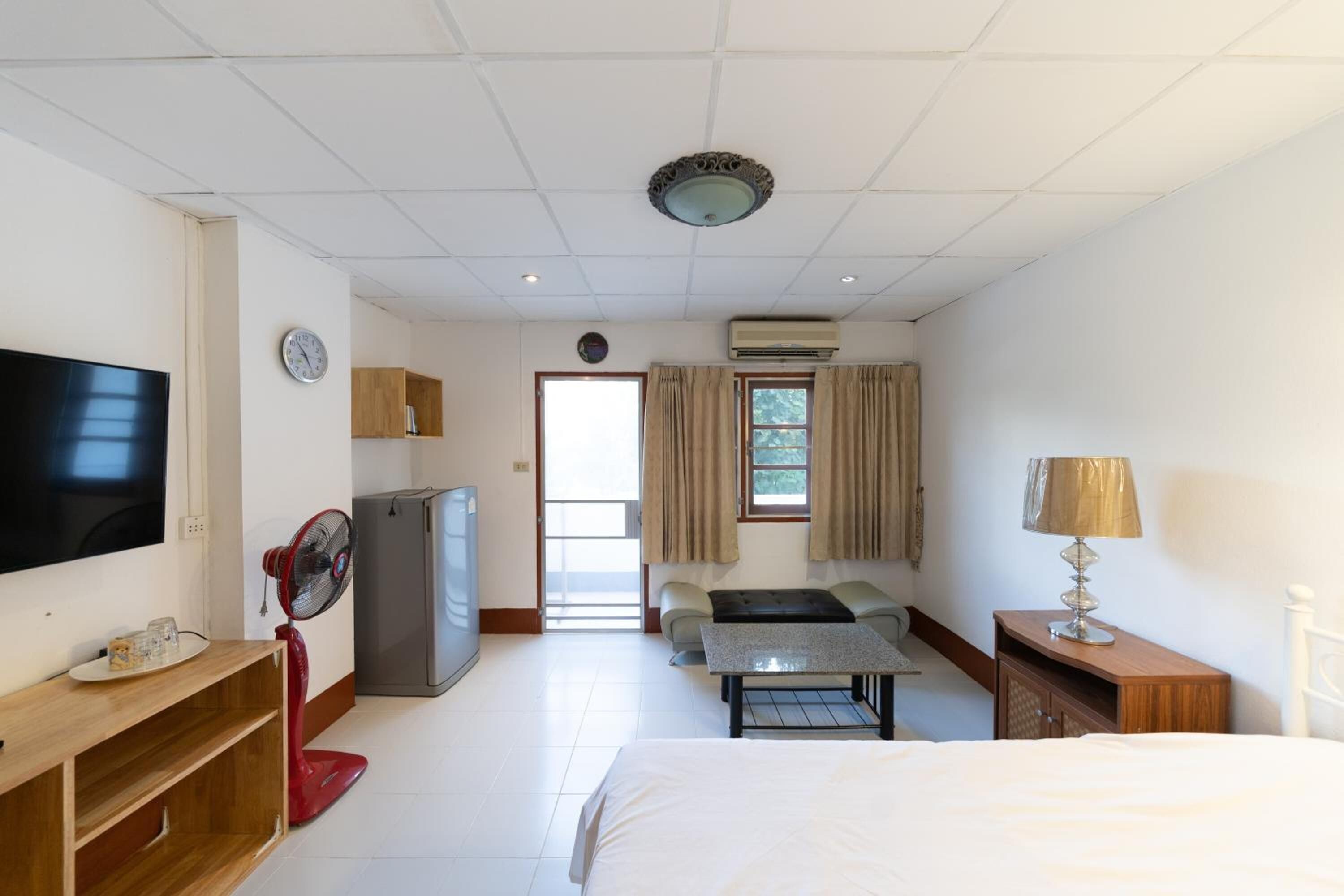 A hotel room with a bed, a TV, a fan, a refrigerator, a table, a lamp, and a window with curtains.