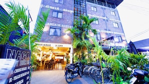 Edificio de ladrillo con letrero que dice 'ANGKOR HOSTEL' y 'GUESTHOUSE'. Hay bicicletas estacionadas frente al edificio, y un cartel anuncia 'NAGA HAPPY HOUR' con precios de hamburguesas.
