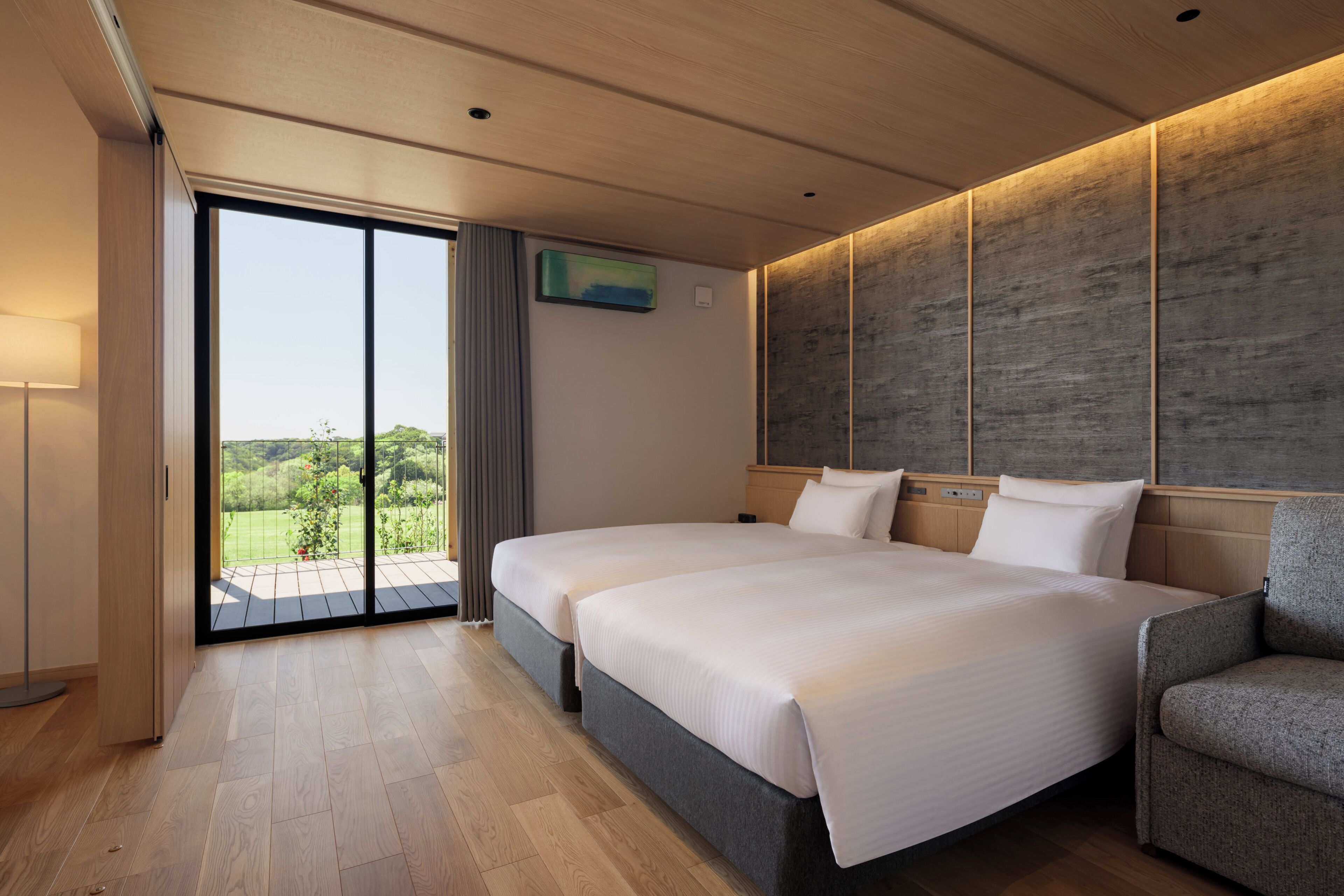 A modern hotel room with two beds, a large sliding glass door to a balcony, and a view of greenery.
