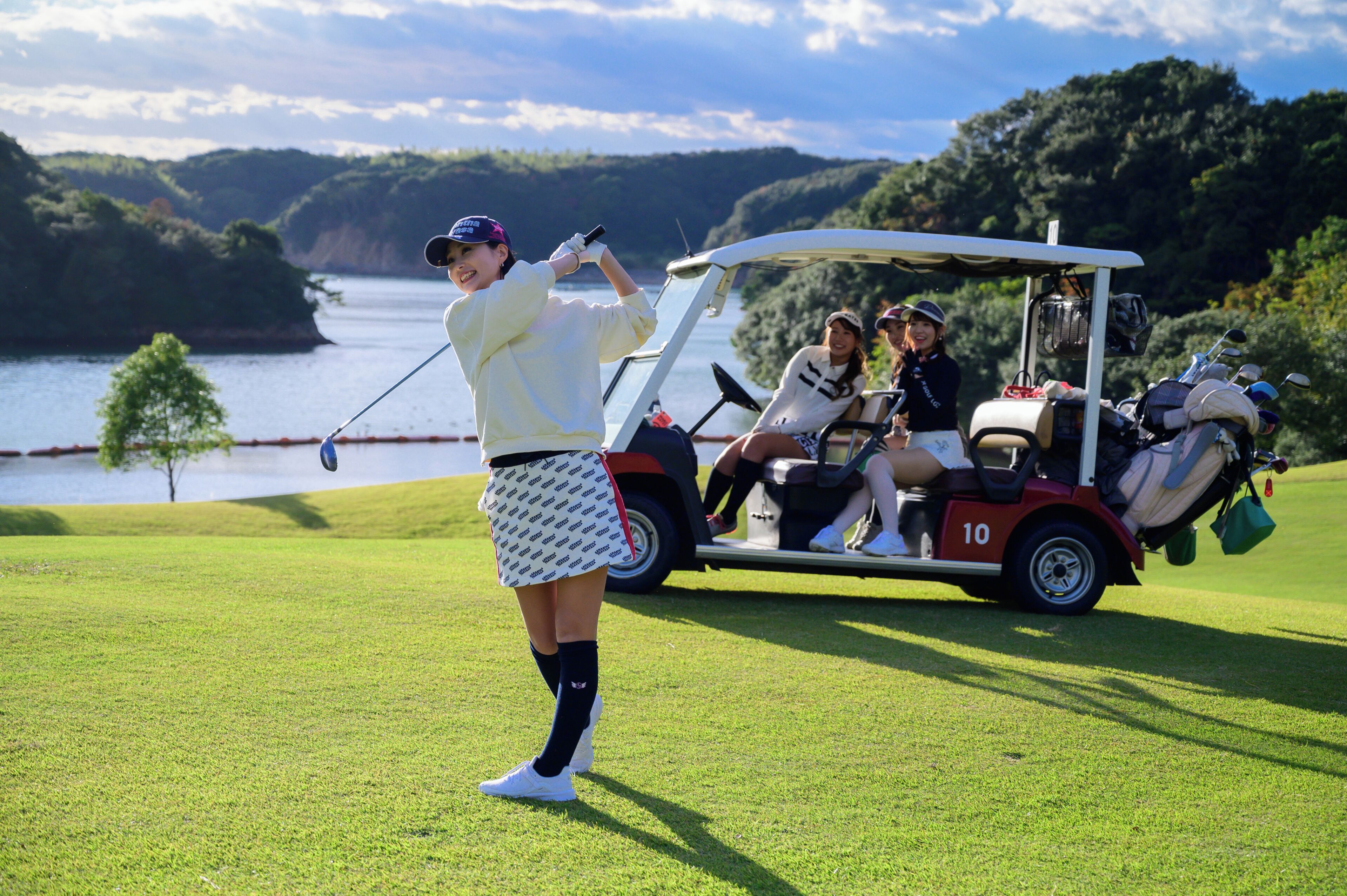 A golf course with a player in a white outfit and a golf cart with passengers.