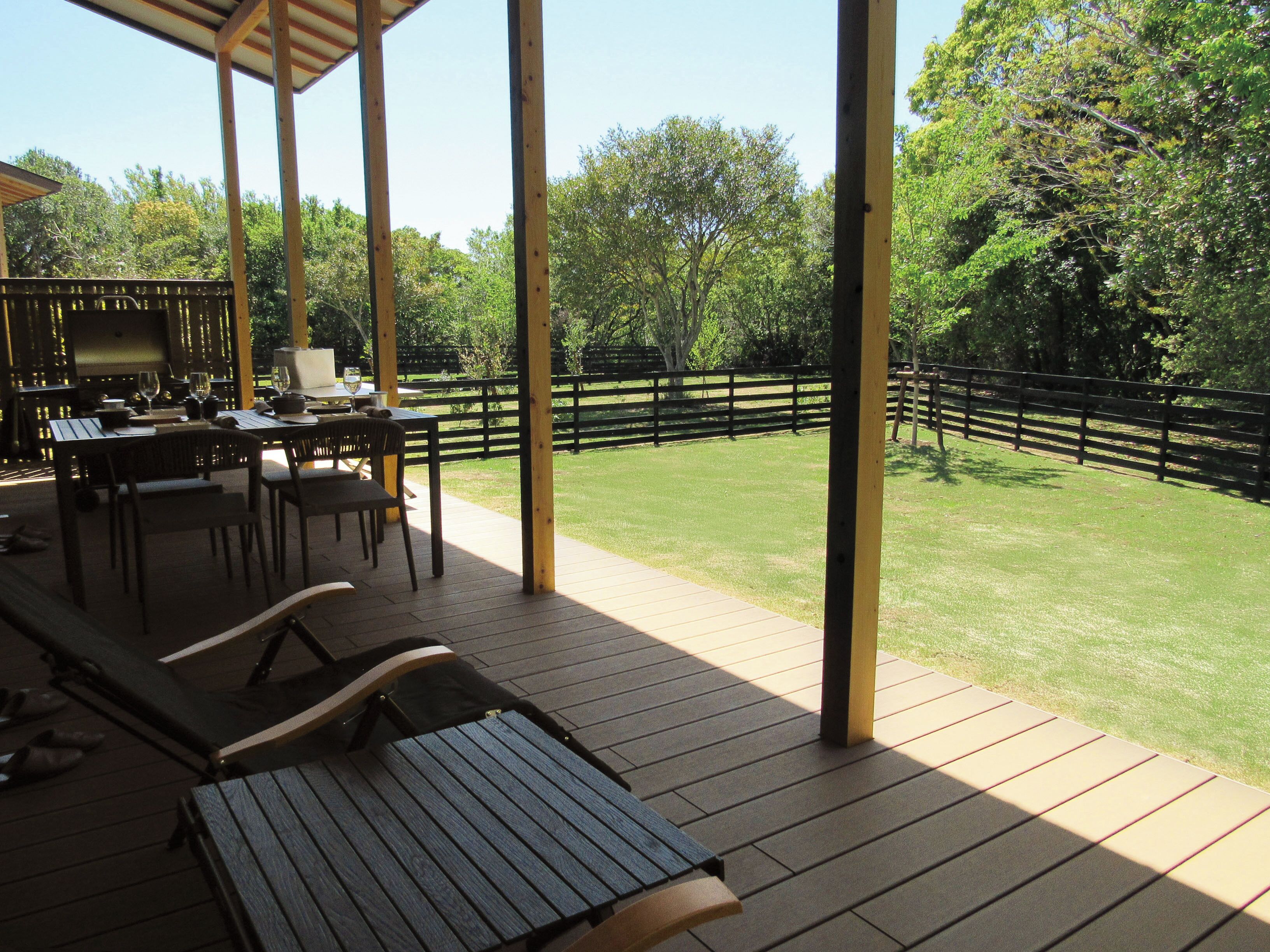 An outdoor patio with tables and chairs, a grill, and a view of a grassy area with a wooden fence.