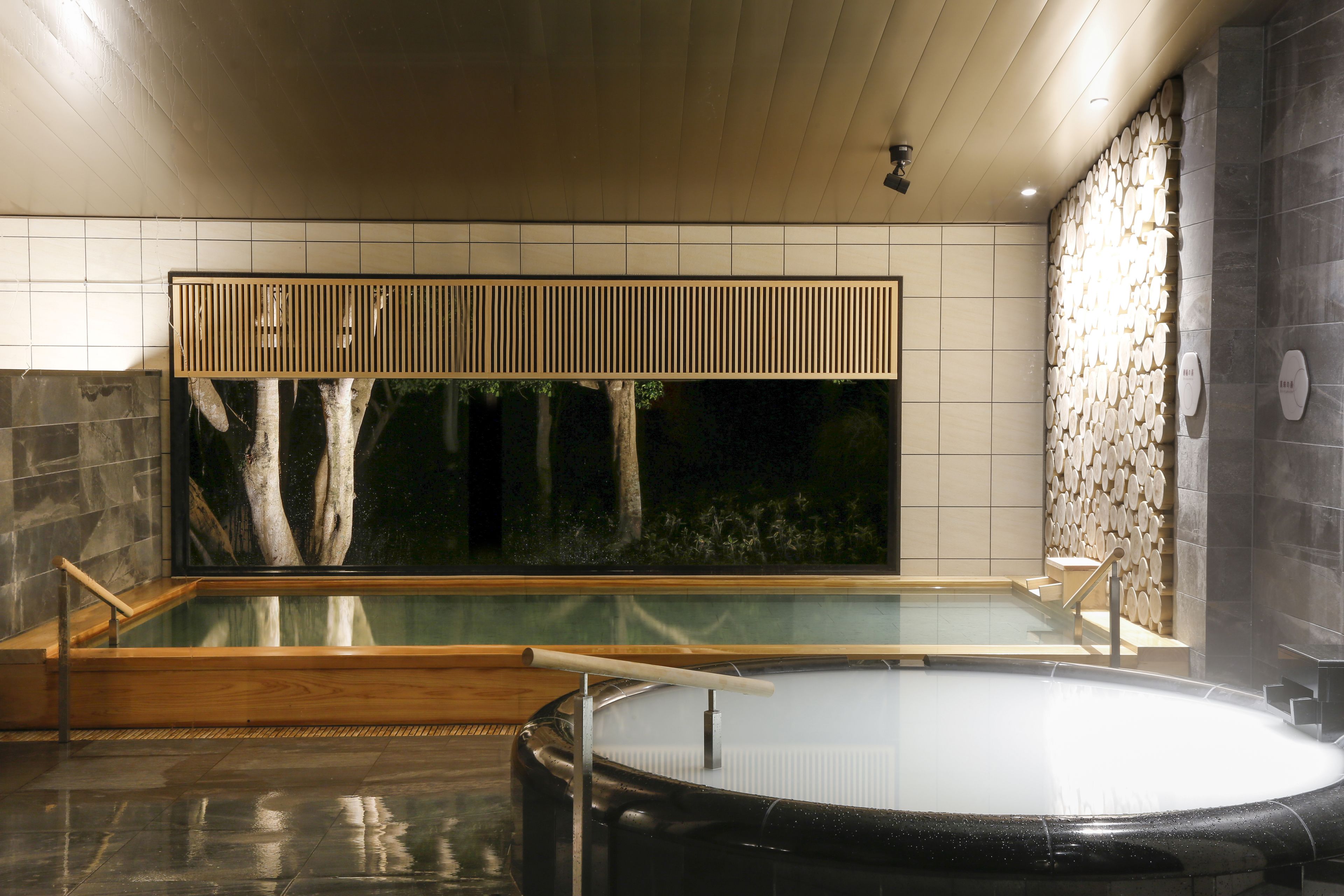 Ett modernt spa med ett stort bubbelbad och en mindre pool, ett tak med träribbor och en stenmur med en vattenfontän.