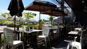 An outdoor seating area with tables and chairs under umbrellas, with a view of the street and buildings in the background. - Coffee Island - Hostel (Koh Samui)