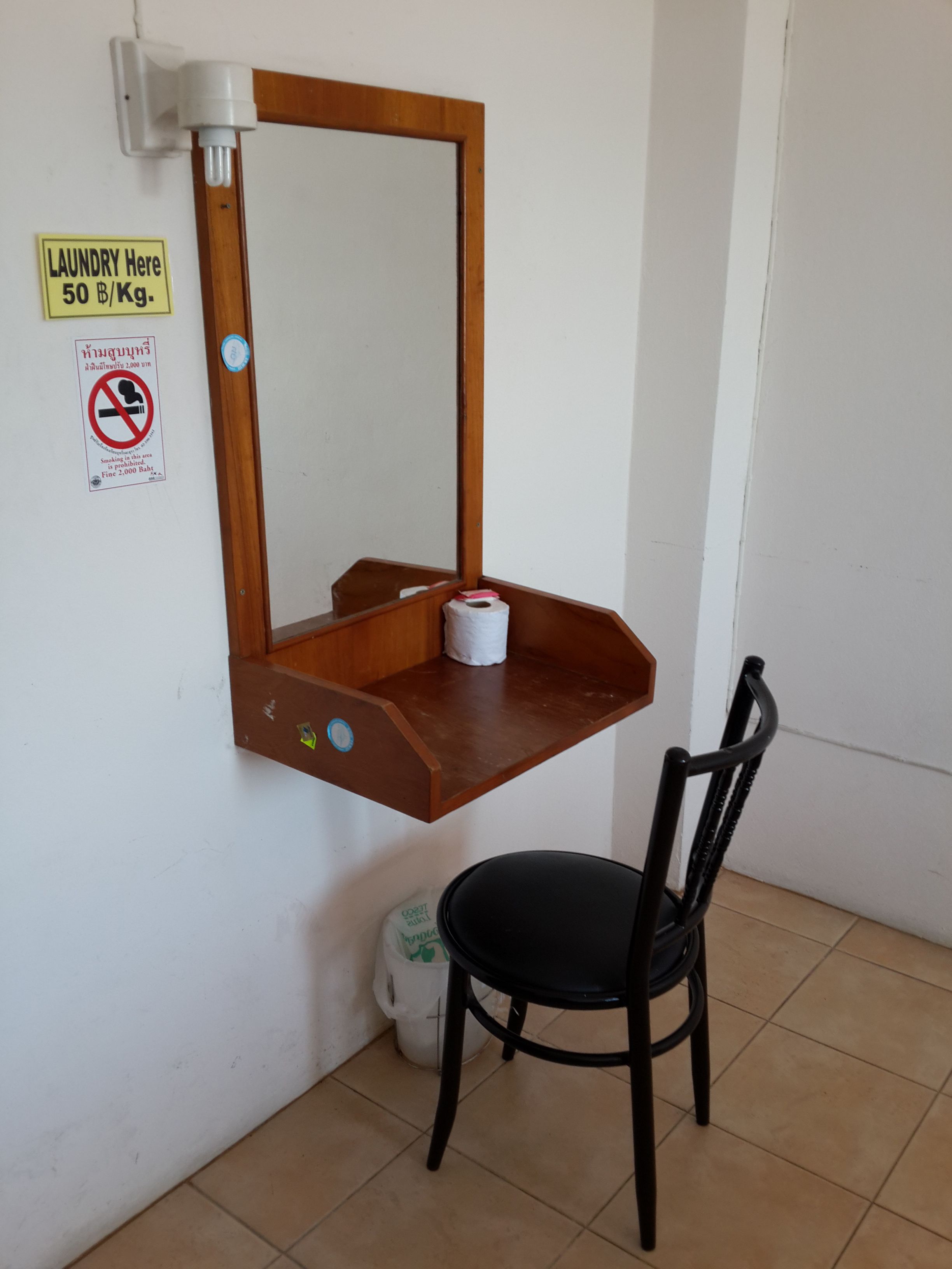 Una habitación pequeña con un escritorio de madera, una silla, un espejo, dispensador de papel higiénico y un cartel que indica los precios de la lavandería.