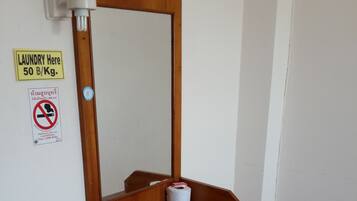 Una habitación pequeña con un escritorio de madera, una silla, un espejo, dispensador de papel higiénico y un cartel que indica los precios de la lavandería.
