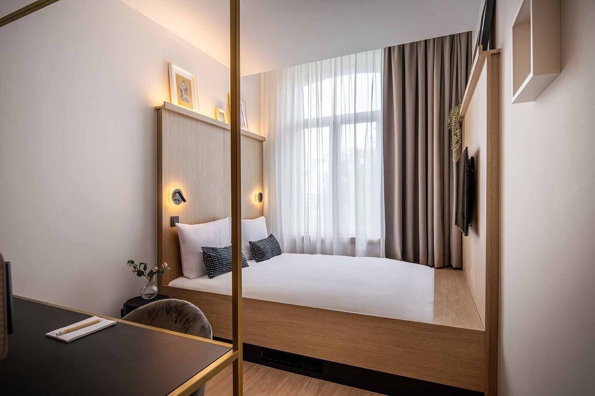 A modern hotel room with a large bed, bedside lamps, a flat-screen TV, and a large window with curtains.